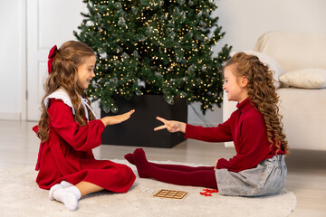 Two little girls friends play rock paper scissors hand game near Christmas tree at home. Children...