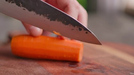 Close-up of a hand using a knife to slice a peeled carrot in half. The vibrant orange vegetable is on a wooden board. The process of preparing fresh ingredients for healthy cooking - Powered by Adobe