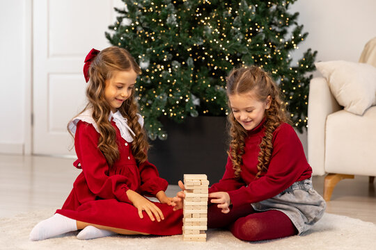 Happy family, little sister girls or best friends are sitting on floor near Xmas tree, playing board game with wooden tower together at home while spending leisure Christmas holidays. - Powered by Adobe