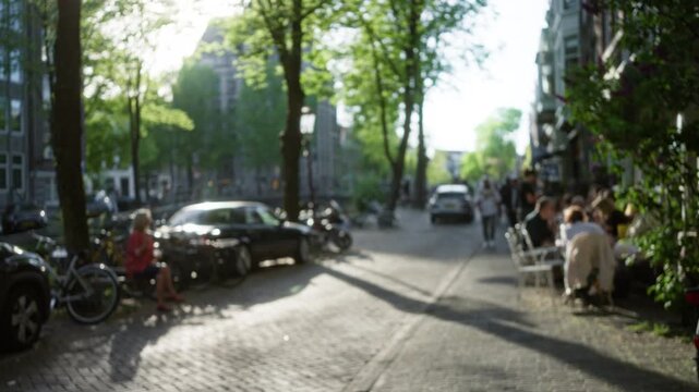 Blurred street scene in a european city with people dining outdoors, trees lining the pathway, and cars parked along the cobbled road.