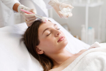 Naklejka premium Anti-aging skincare. Cosmetologist applying clay mask on face of young lady lying on table, enjoying beauty treatments in spa salon