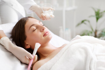 Young woman enjoying facial mask treatment at beauty salon, cosmetologist applying mask on female...