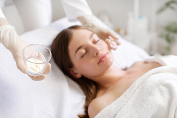 Focus on bowl with mask, beautician applying face mask to rejuvenate lady skin, treatment of...