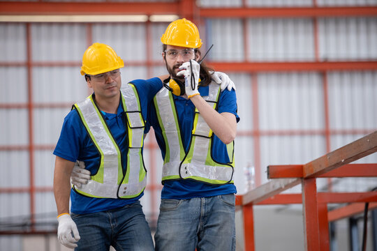 Construction worker supports injured colleague while calling for emergency help in warehouse. Workplace injury recovery, teamwork, and industrial safety response concept.
