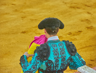 A matador in an ornate turquoise jacket and traditional black hat holds a bright yellow and pink...