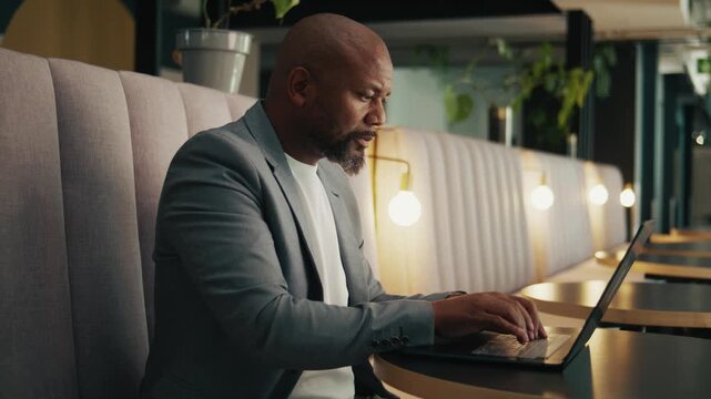 A serious mature black businessman in blazer using laptop in office lounge
