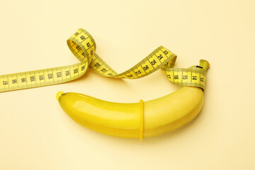 Sex education. Banana with condom and measuring tape on beige background, top view
