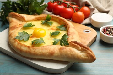 Tasty khachapuri with cheese, egg, parsley, tomatoes and spices on light blue wooden table, closeup