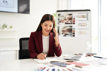 Concentrated young interior designer woman sitting at table with blueprint drawing ,swatches and material , discussing interior design with client  by phone, choosing materials for decorations.