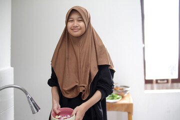 Asian muslim woman smiling while holding bowl of shallots, cooking in the kitchen