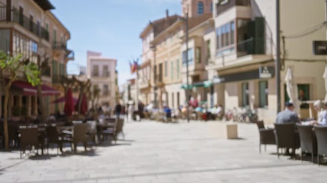 Blurry scene of a mediterranean old town street with people walking, quaint outdoor cafes, and historical architecture under a clear blue sky.