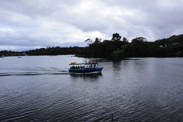 Fototapeta premium Scenic Boat Ride on Calm Lake with Tourists Enjoying Nature and Tranquil Waters, Surrounded by Green Hills, Forest, and Cloudy Sky – Perfect for Travel, Adventure, and Relaxation Concepts