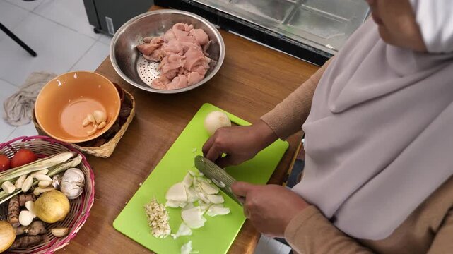 Asian muslim woman peeling, cutting and slicing onion and garlic. Preparing marinade for chicken meat