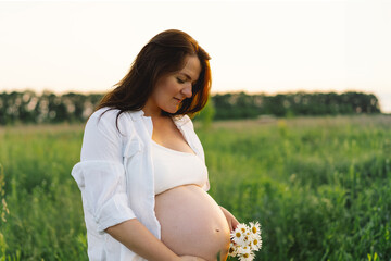 A pregnant woman stands in a green field. She gazes lovingly at her baby bump as the sun sets...