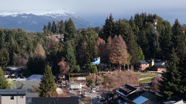 Drone push-in over Villa La Angostura, showing the Argentine flag waving at the civic monument along Avenida Arrayanes, framed by dense Patagonian forest and snow-capped Andes peaks beyond.