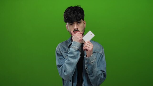 Young man standing against a vibrant green background, holding a card and wearing a denim jacket, thoughtfully posing with a finger on his chin and eyes closed.