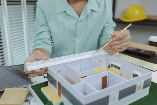 Asian male architect sitting at a desk, measuring a modern house model with triangular ruler and pencil. Office setup includes design tools, blueprints, and safety gear, suggesting architectural work.