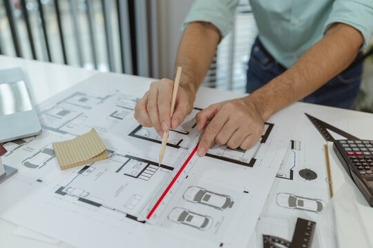 An architect using a pencil and a scale ruler to measure house floor plans. Various design tools and model pieces are on the desk, indicating active architectural planning process.