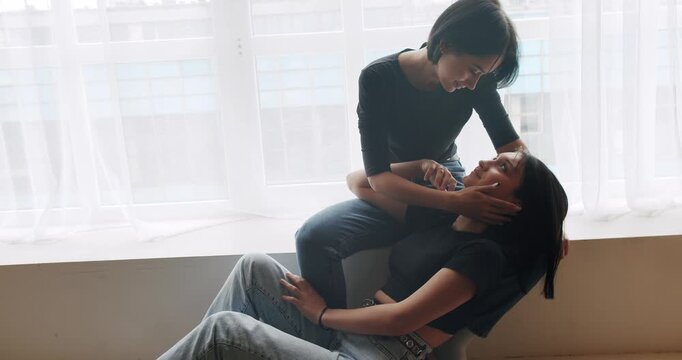 Low angle handheld shot of young lesbian couple smiling and touching hands gently while sitting on sofa against window in light living room at home. Lifestyle and relationships. LGBT concept