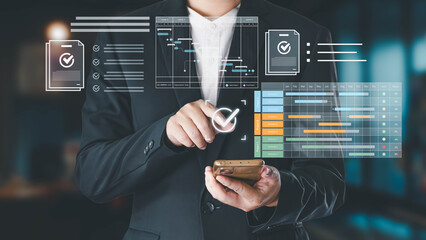 A businessman in a suit uses his smartphone to interact with a futuristic digital interface displaying project management tools, including Gantt charts and checklists, in a modern office environment.