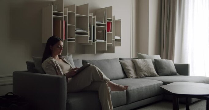 Woman reading a book while seated on a comfortable gray couch in a modern, well-designed living room with shelves and soft lighting. Woman Reading Book on Comfortable Couch in Modern Living Room