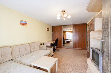 Cozy living space featuring a beige sofa, light wood table, desk, office chair, mirrored closet, and a vintage TV stand. Room has yellow walls and textured carpet