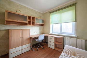 cozy room with wooden furniture: wardrobe, desk, shelves. A rolling blind covers the window, illuminating the space with soft light
