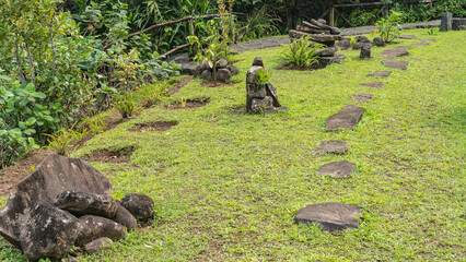 A pedestrian path made of flat stone sections runs through the green lawn in the park. Pyramids of mossy boulders on the grass. Ferns, tropical plants are growing. Mauritius. Landscape design.