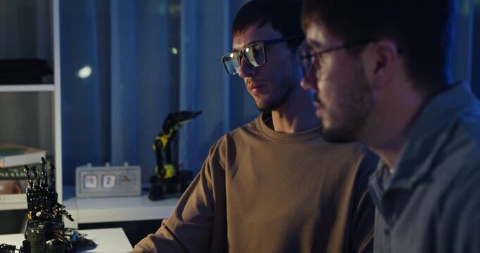 Team adult male engineer collaborate on robotic hand project using AI code solutions machine learning at tech workspace night. Artificial intelligence interface, collaboration prototype development.