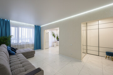 bright living room with white marble floors, a gray sofa with blue pillows, blue curtains, and recessed lighting. An opening leads to a kitchen