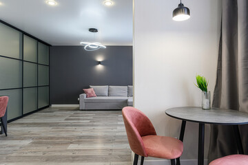 Modern interior featuring a pink chair, round table, and glimpse into a living room with a grey sofa and decorative lighting. Minimalist design