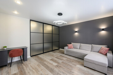 Cozy living room featuring a gray sofa with pink pillows, a desk with a chair, and a frosted glass partition. Modern lighting and wood flooring enhance the space