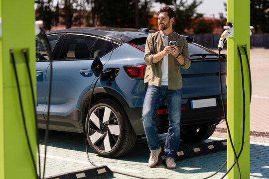 Man using smartphone while charging electric car at charging station