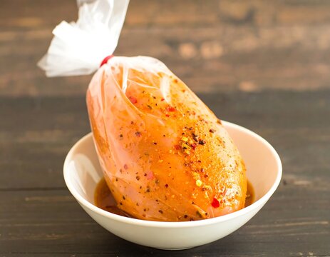 Marinated poultry breast in a plastic bag, resting in a shallow white bowl on a wooden surface