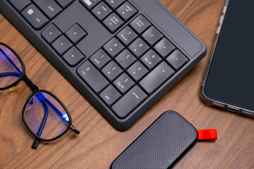Modern workspace with keyboard, smartphone, hard drive, and blue-lens glasses on wood desk—clean tech setup for digital productivity and device ads.