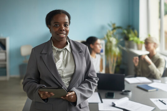 Portrait of young adult Black woman standing in modern office holding digital tablet, smiling at camera while diverse colleagues working in background during business meeting