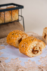 Two pieces of savory bread rolls sprinkled with sesame seeds, showing a visible filling inside, placed on a lace-patterned cloth with a bread basket in the background.