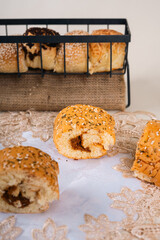 A close up shot of golden brown bread rolls with sesame seeds and herbs on top, placed on a lace cloth with a basket of assorted bread in the background.