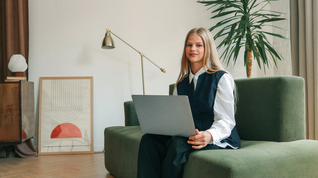 Young woman sitting on green sofa with laptop, smiling and working in stylish retro-inspired interior with modern lamp, framed art poster, and indoor plant