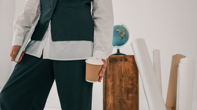 Student in black vest and white shirt holding a coffee cup and laptop, standing near wooden pedestal with globe and rolled paper sheets in studio