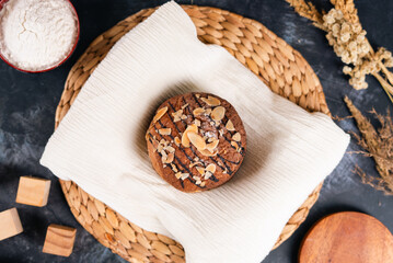 A round chocolate bread topped with sliced almonds and drizzled chocolate, placed on a white cloth over a woven mat with baking ingredients around.