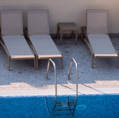 Empty beige sun loungers and table by swimming pool with metal ladder, outdoor relaxation area in...