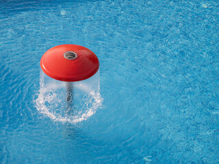 Red mushroom-shaped fountain in swimming pool with flowing water creating ripples on blue surface.