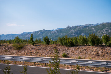 Naklejka premium Modern highway with guardrails leading through rocky landscape and green mountains under blue sky. Scenic transportation route in Croatia.