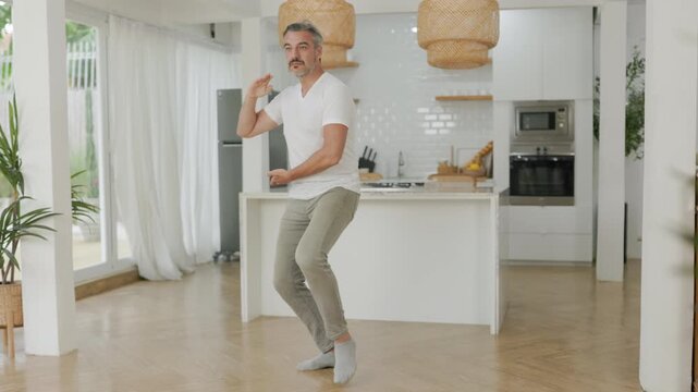 Mature man practicing Tai Chi in movement indoors, standing in a bright kitchen with a relaxed and focused expression.