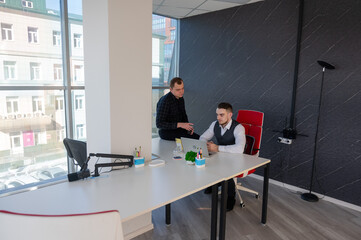 Caucasian man standing at colleague's desk discussing work. 
