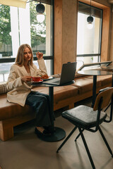 Young woman in beige blazer sitting at cafй table with laptop, drinking coffee and enjoying breakfast with croissant and salad, modern interior with large windows