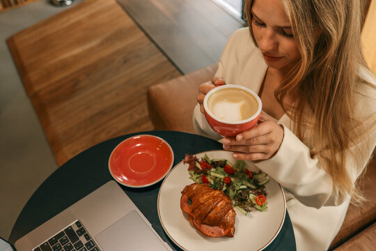 Woman enjoying coffee with croissant and fresh salad at cozy cafe table, holding red cup and smiling, breakfast or brunch concept - Powered by Adobe