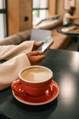 Close-up of red coffee cup with latte on table, blurred background shows woman using smartphone, cozy cafe atmosphere