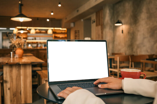Person using laptop with blank screen at cafe table, red coffee cup nearby, cozy interior with warm lighting and wooden furniture